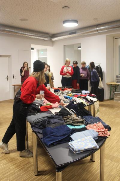 Mehrere Frauen stehen in einem großen Raum um einen Tisch. Auf dem Tisch befinden sich Kleiderstücke in verschiedenen Formen und Farben. Manche von den Frauen reden miteinander, andere schauen sich die Kleidung an. 