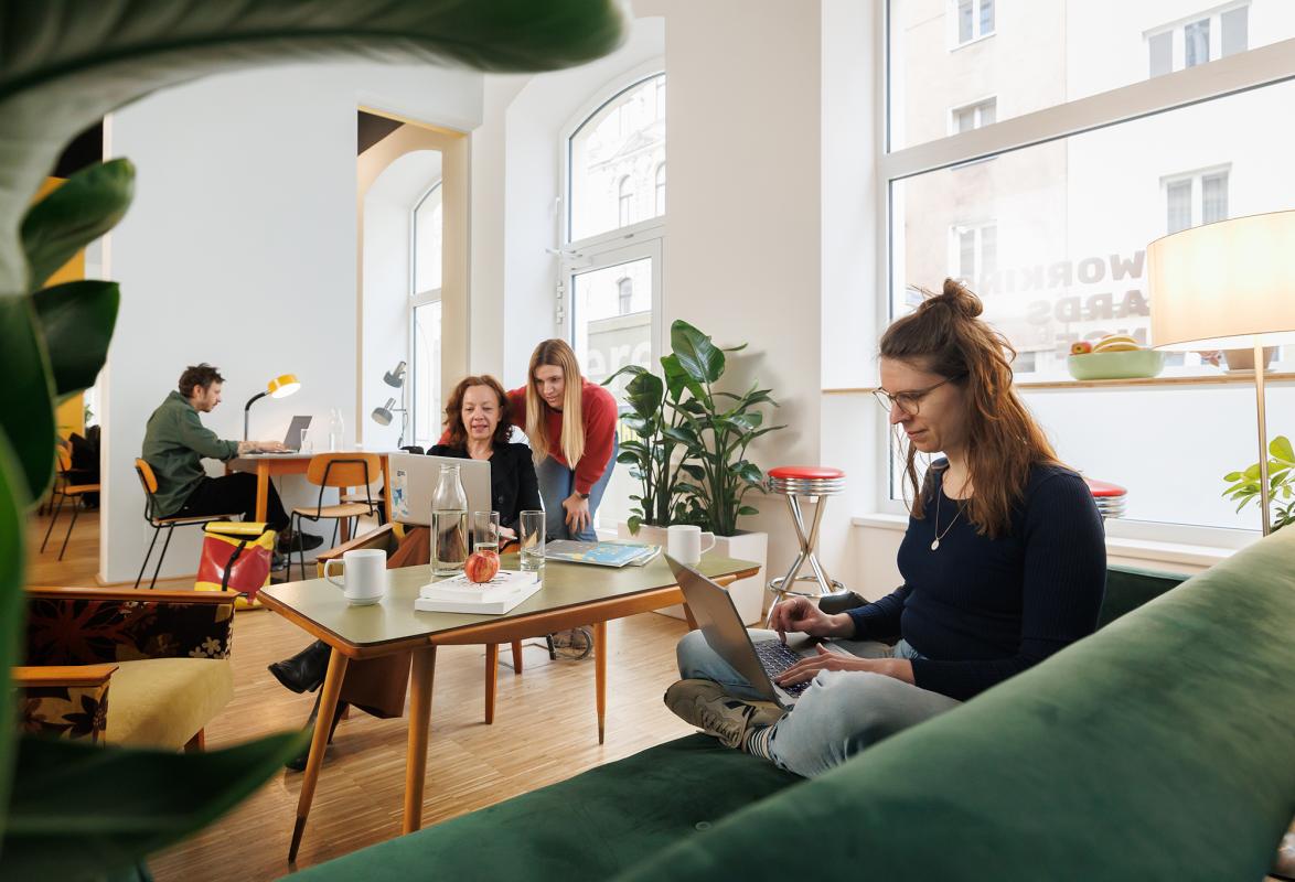 Im Vordergrund sitzt eine junge Frau auf einem Sofa und arbeitet am Laptop. Im Hintergrund sieht man zwei Frauen die etwas an einem Laptop besprechen und einen Mann der intensiv an einem Laptop arbeitet. Sie sitzen im Coworking-Space.