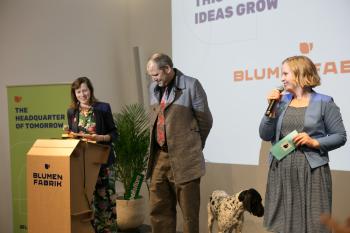 Agnes Zauner steht auf der Bühne und spricht in ein Mikrophone, das sie in der Hand hält. Neben ihr steht Rene Fischer und die Moderatorin Eva Gänsdorfer 