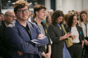 Im Fokus steht Nikolai Miron, der im Publikum steht, aber in die Kamera lächelt. Im Hintergrund sieht man weitere Personen aus dem Publikum