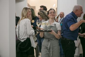 Eine Kellnerin mit grauer Schürze schaut in die Kamera und lächelt. Sie hält ein Tabeltt mit Weißwein-Gläser in der Hand.
