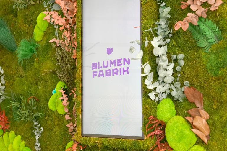Trockenblumen-Wand mit Moos in Detail-Ansicht. In der Mitte befindet sich ein Bildschirm worauf das Logo der Blumenfabrik zu sehen ist. 