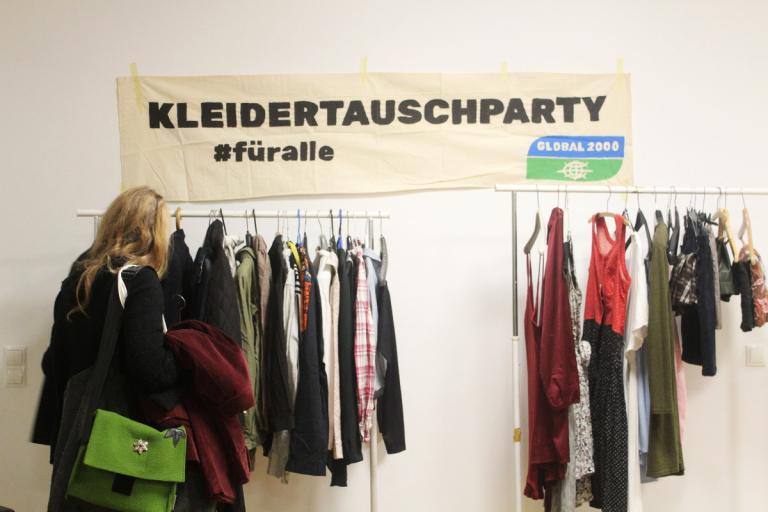 Frau begutachtet bei einer Kleiderstange verschiedene Kleidungsstücke. Über der Kleiderstange befindet sich ein Bunner worauf steht "Kleidertauschparty #füralle"