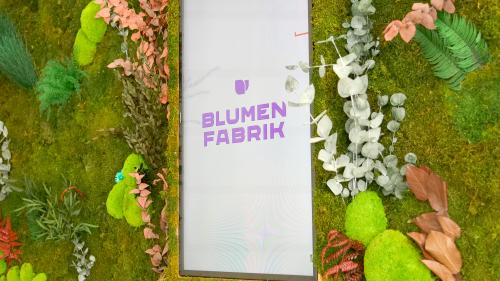 Trockenblumen-Wand mit Moos in Detail-Ansicht. In der Mitte befindet sich ein Bildschirm worauf das Logo der Blumenfabrik zu sehen ist. 